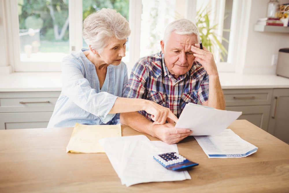 An elderly couple sitting at a table, reviewing financial documents and bills, emphasizing the challenges in managing finances and estate planning for aging parents during the COVID-19 pandemic.