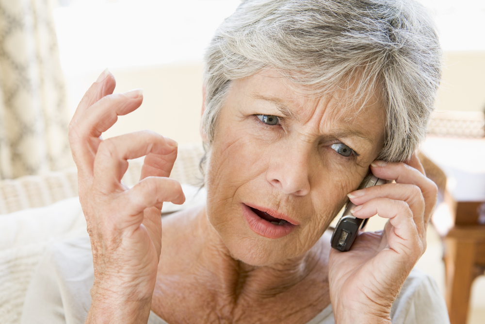 An elderly woman appears irritated while holding a phone, illustrating the frustration faced by seniors during Medicare enrollment due to technical difficulties with Social Security Administration phone lines.