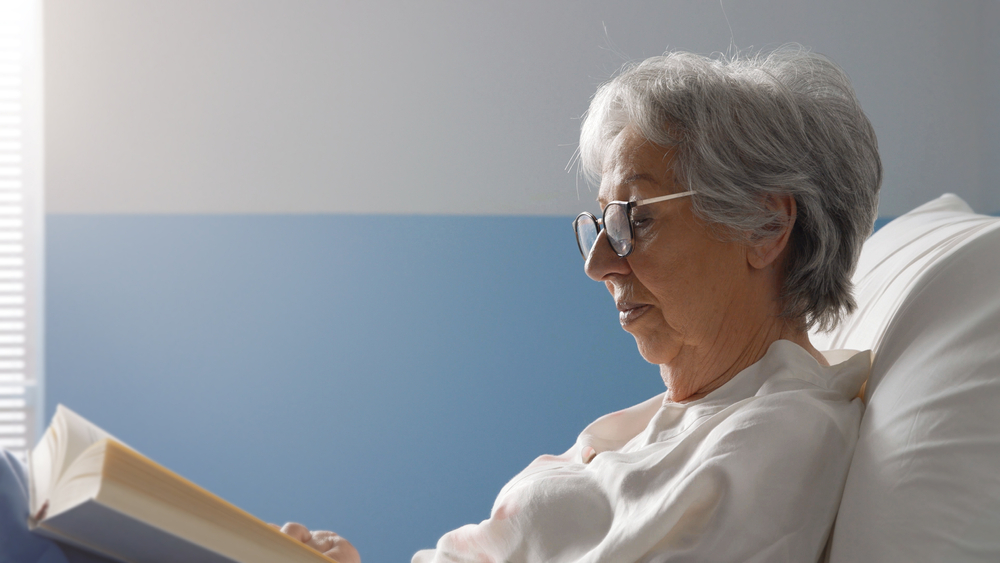 Elderly person in a nursing home bed, reading a book, highlighting the management of personal needs allowance under Medicaid.