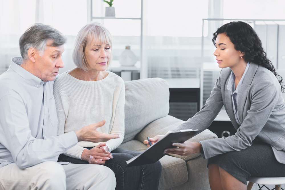 Senior man and his wife sitting on a couch consulting with a female lawyer about estate planning documents, emphasizing their concern for future planning in elder law.