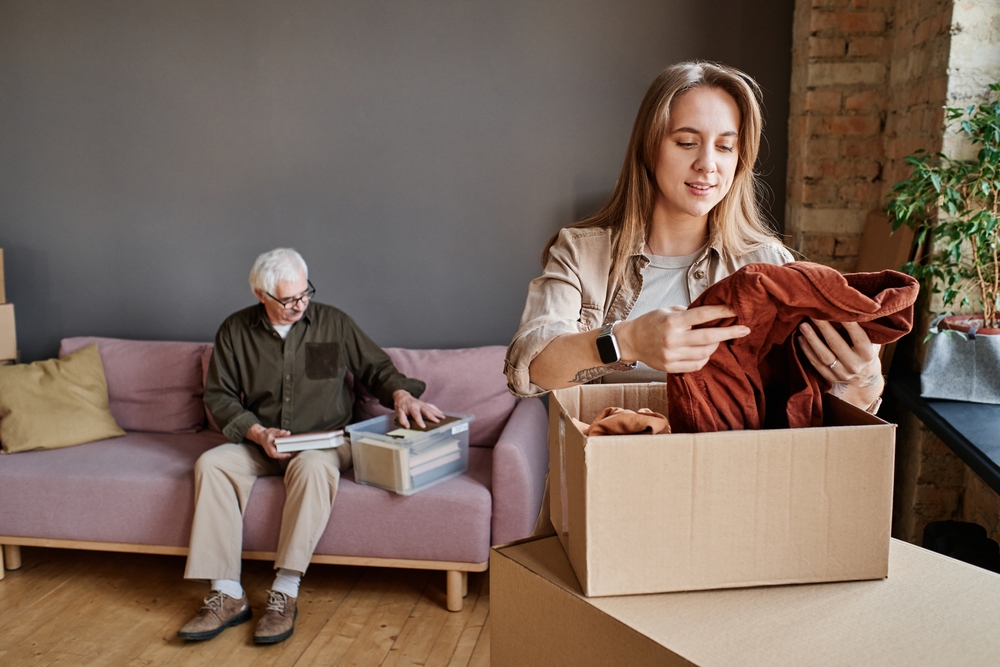 A young woman packs clothes for her grandfather in an apartment, symbolizing the preparation involved in transferring guardianship across state lines within the context of estate planning and elder law.