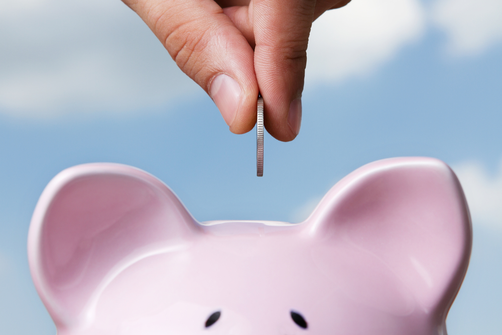 A male hand places a coin into a pink piggy bank, symbolizing increased retirement contributions and estate planning strategies for 2023.