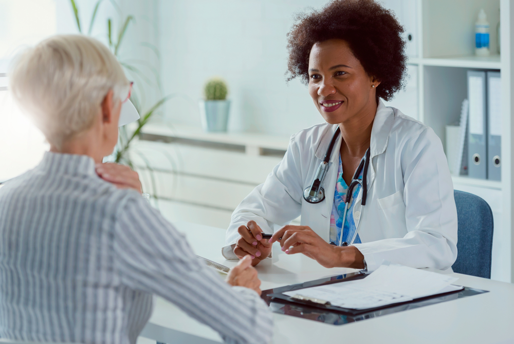 An African American female doctor sitting in her office, talking with a senior female patient, discussing the importance of getting a second opinion and Medicare's role in covering medically necessary procedures.