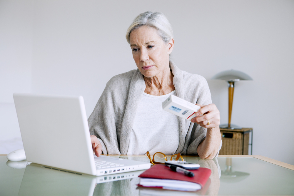 An elderly woman sitting at a table, using a laptop to purchase medications online, emphasizing the affordability of generic drugs through online pharmacies in the context of estate planning and elder law.