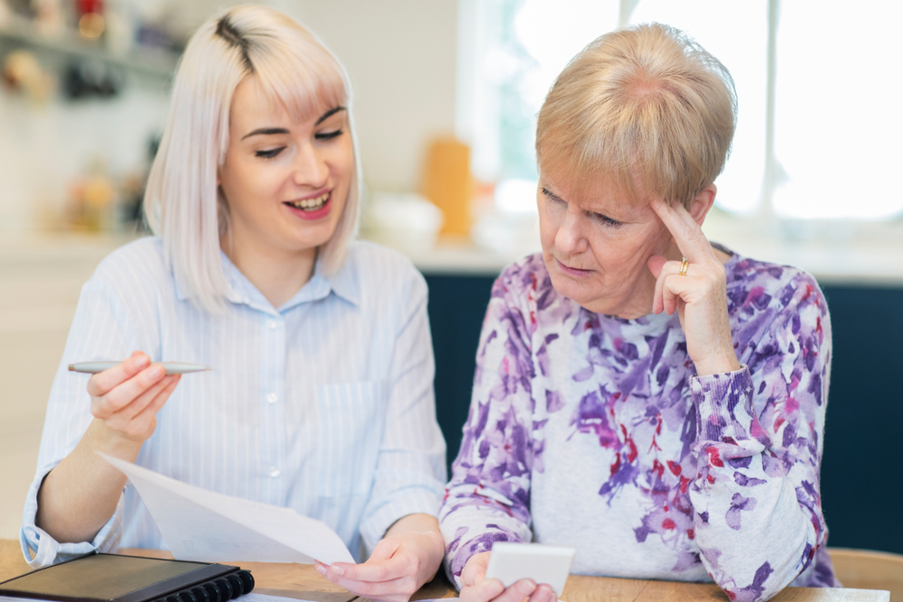 A woman helps an elderly neighbor with financial paperwork, emphasizing the need for financial guardianship and elder law planning to protect vulnerable seniors in managing their finances and estate.