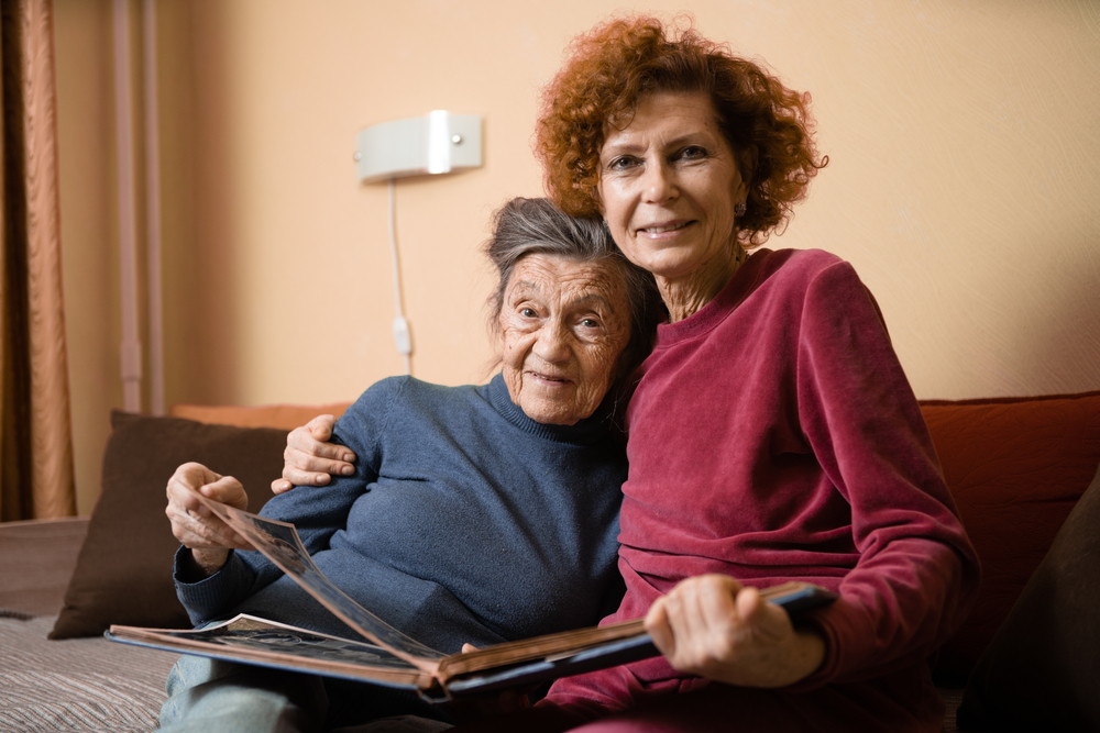 Senior woman and her adult daughter looking at a photo album together on a couch in the living room, symbolizing the need for discussions about estate planning and the impact of Medicaid eligibility requirements.