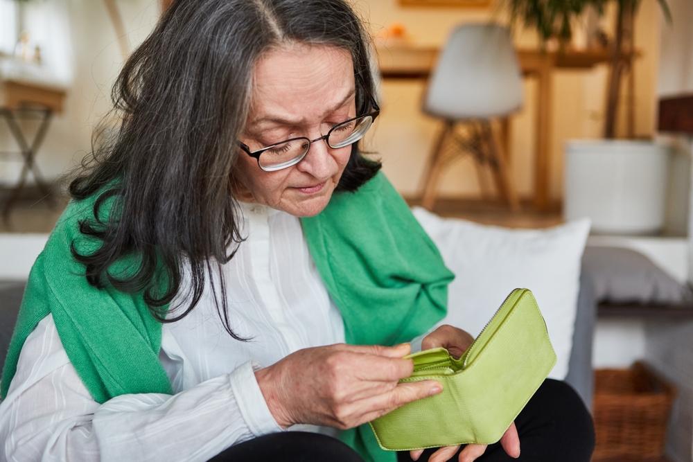 An elderly woman sits at home, looking worried as she examines her empty wallet, representing the economic challenges many seniors face and the consideration of selling life insurance policies to cover expenses.