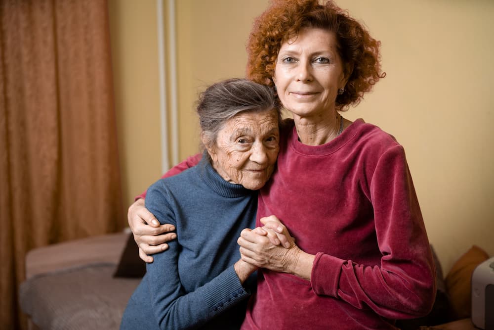 Elderly woman with dementia smiles with her middle-aged daughter at home, illustrating the emotional and financial challenges of family caregiving in the context of proposed legislation for cost relief.