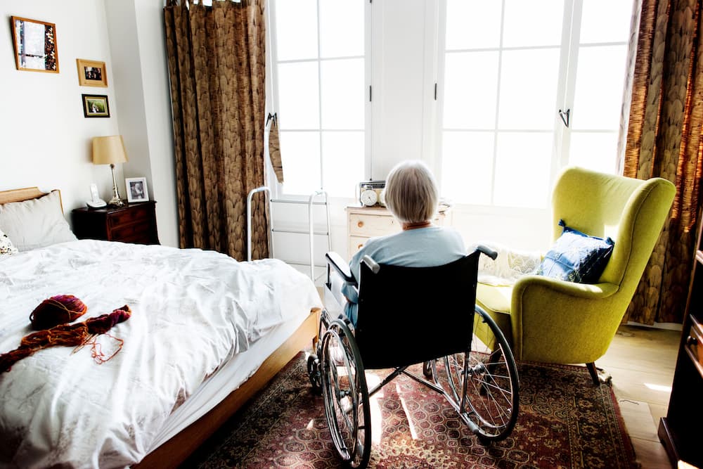 A senior woman in a wheelchair sits alone in a sunlit bedroom, facing away, representing the housing affordability and support challenges faced by older adults in the context of increasing longevity and the elder law implications of the housing crisis.