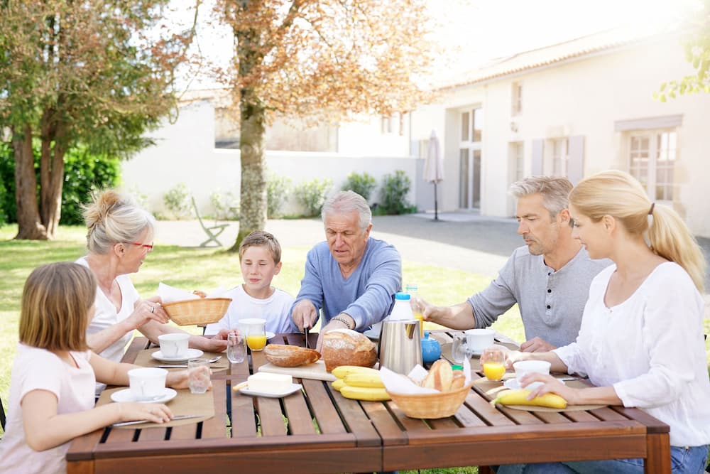 A multigenerational family has breakfast in the backyard of a modern home, highlighting estate planning and Medicaid strategies to preserve family assets.