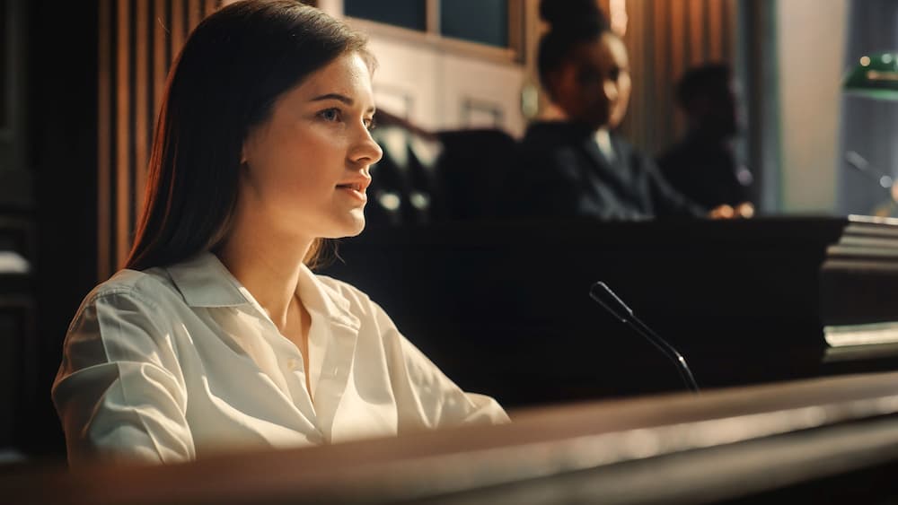 A female witness answers prosecutor questions in a probate court setting, with a judge listening in the background, representing disputes that may arise in estate planning and elder law matters.