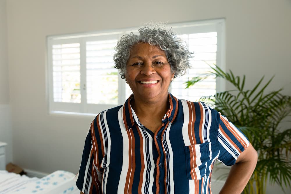 An elderly African American woman stands smiling in her bedroom, illustrating the concept of aging in place supported by HUD housing programs.