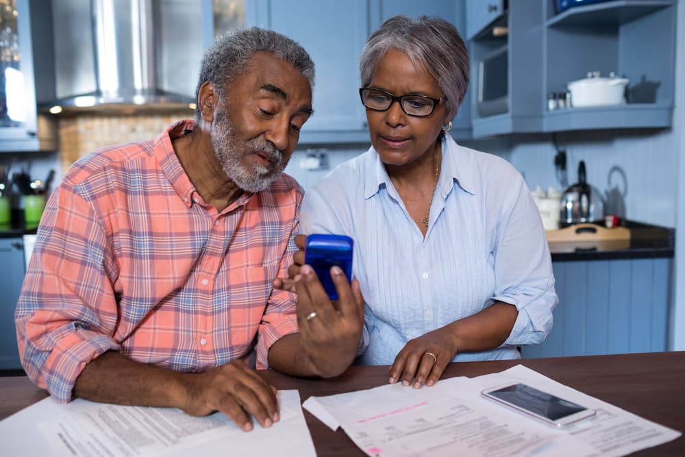 An older couple sitting in their kitchen, using a calculator and examining financial and retirement paperwork, emphasizing the significance of financial literacy in retirement planning for seniors.