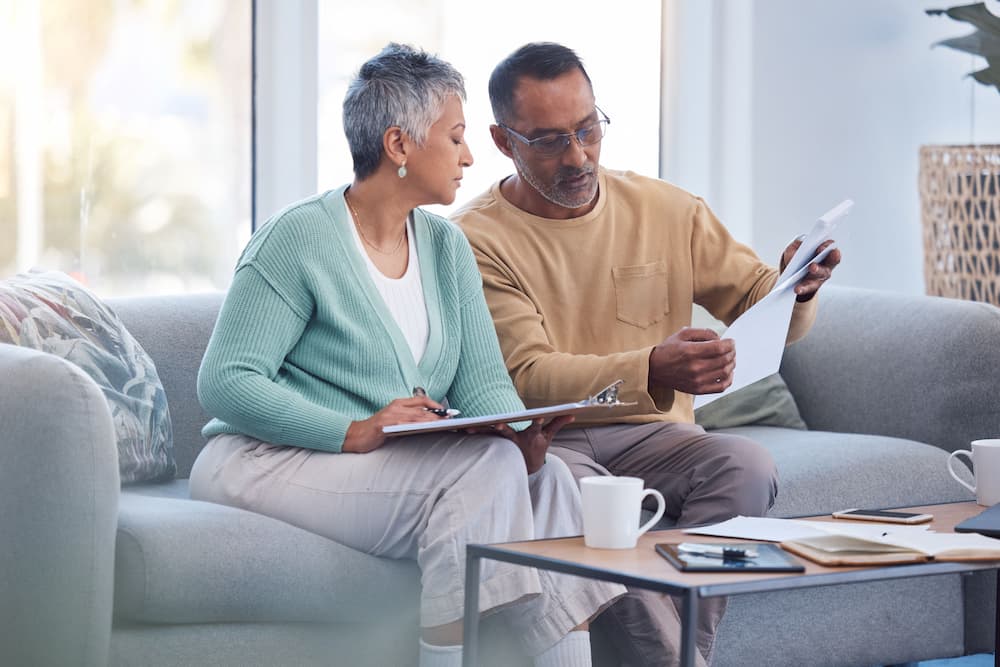 An elderly couple seated on a sofa looks through financial papers and documents, symbolizing the critical role of estate planning and elder law in preparing for the high costs of long-term care.