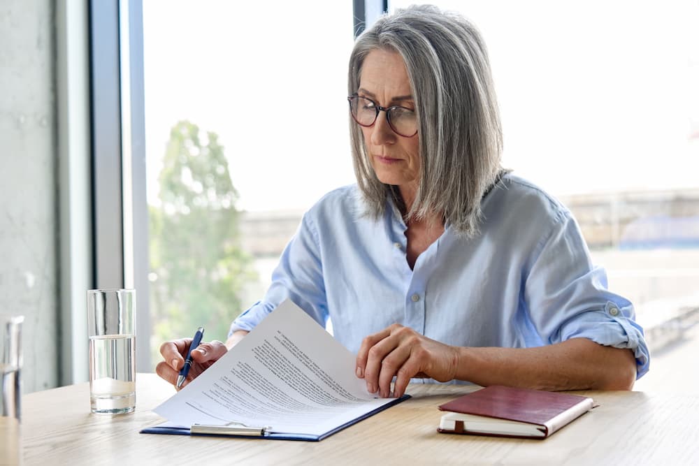An elderly woman sits at a desk in an office, signing a legal document, illustrating the role of a durable power of attorney in estate planning and avoiding guardianship.