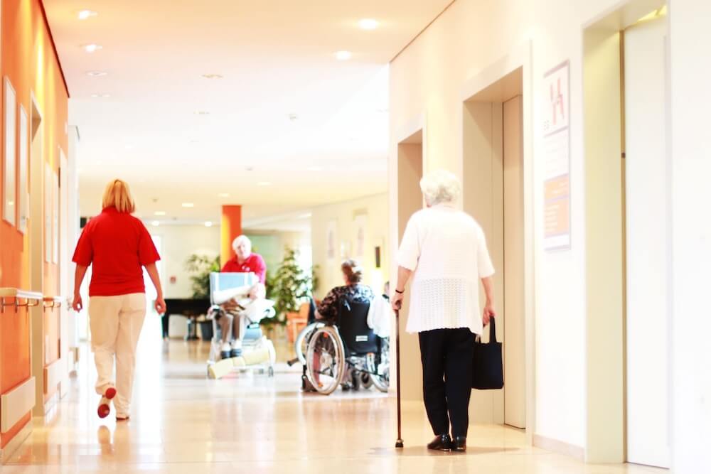 An older woman with a cane and other senior residents in wheelchairs moving through a well-lit corridor as staff in red shirts assist them.
