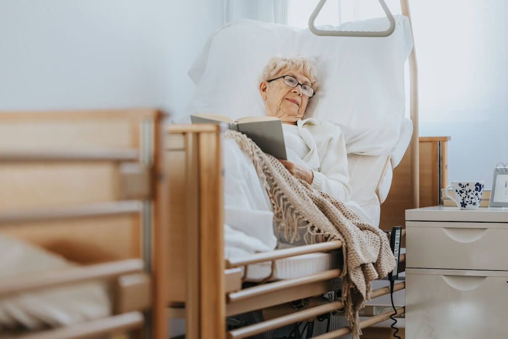 Elderly woman wearing glasses and reading a book in a nursing home bed, with bed rails and a bedside table visible in the room.