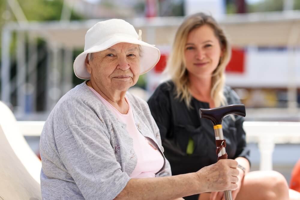 Elderly woman with white sunhat holding a cane, sitting beside a younger woman outdoors.