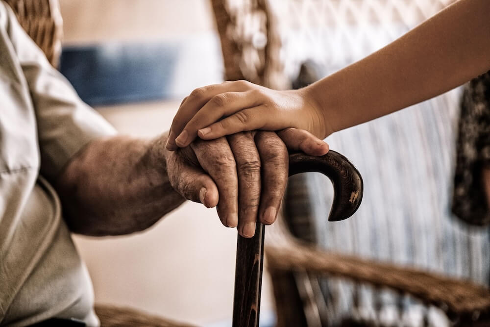 Young hand gently resting on an elderly person’s hand holding a cane, symbolizing care and guardianship.