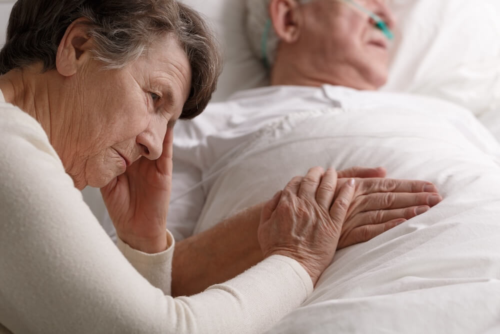 Elderly woman sitting beside her unconscious husband in a hospital bed, gently holding his hand.