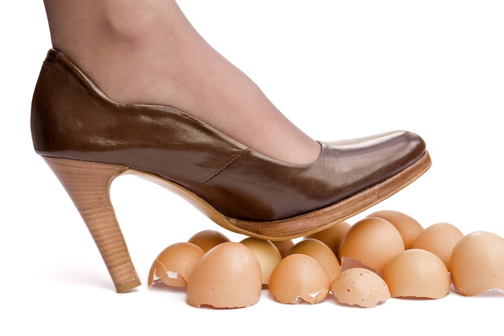 A person stepping on fragile eggshells while wearing a brown high-heeled shoe, symbolizing the risks and instability of living without an estate plan.
