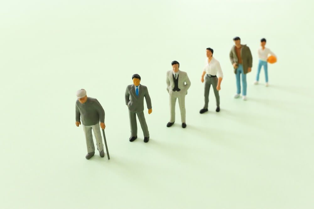 Miniature figurines of men arranged in a line from an elderly man with a cane to a young man holding a basketball, representing different generations.