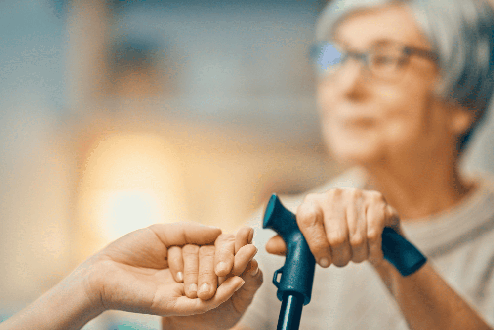 Caregiver holding an older adult’s hand while assisting with a walker in a nursing home setting