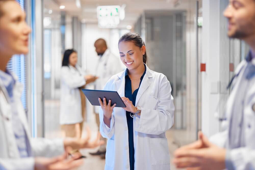 Female physician in white coat reviewing practice management data on a tablet in a hospital corridor